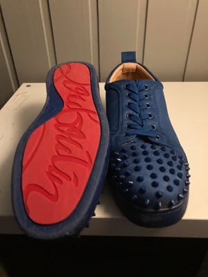 Blå Christian Louboutin sneakers - Säljer ett par blå sneakers från Christian Louboutin med ikonisk röd sula och coola nitar på tån. Skorna har snörning och är tillverkade i mocka med skinnfoder. Perfekt för dig som vill sticka ut med en edgy och lyxig stil.