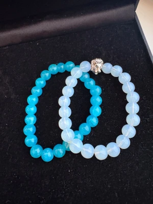 Två pärlarmband i blått och vitt - Säljer två snygga pärlarmband, ett med klara turkosa pärlor och ett med skimrande vita pärlor. Det vita armbandet har en cool detalj med ett lejonhuvud i silverfärg. Perfekt för dig som gillar att mixa och matcha accessoarer.