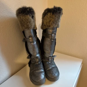 Silvriga boots med päls från Kioss - Snygga silvriga boots från Kioss Fashion med fluffig fuskpäls upptill och dekorativa spännen. Skorna har en robust platt sula och dragkedja baktill. Perfekta för att sticka ut under kalla dagar med sin glittriga finish och coola detaljer.skorna är väldigt varma man kan ha de ute när det är minus grader/snö.orginal pris 4000/3000 
