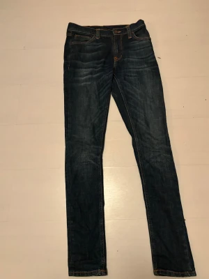 Mörkblå skinny jeans från Nudie Jeans - Snygga mörkblå jeans från Nudie Jeans med klassisk femficksdesign och kontrastsömmar i orange. Modellen är skinny med smal passform och raka ben. Jeansen har normal midja och är tillverkade i slitstarkt denimtyg av bomull.. Nästan oanvända och skicket är helt otroligt. Detta är ett kap nu till jul!! W29 L32