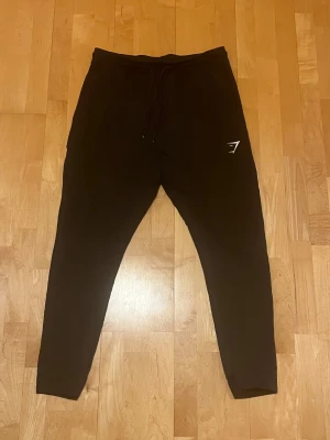 Svarta mjukisbyxor från Gymshark strl M - Svarta mjukisbyxor från Gymshark i storlek M. De har en slim passform, snörning i midjan och en vit Gymshark-logga på vänster lår samt två fickor med dragkedja. Materialet är mjukt och skönt, perfekt för vardag eller träning. 
