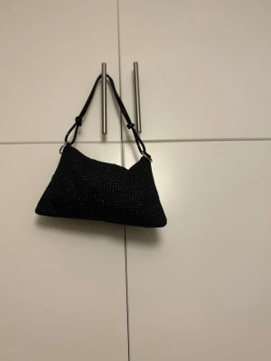 Svart glittrig axelväska - Svart axelväska med glittriga detaljer och rektangulär form. Väskan har ett justerbart axelband med knutar och ett coolt mesh-liknande material som ger extra shimmer. Perfekt för att lyfta din outfit med lite extra bling💕