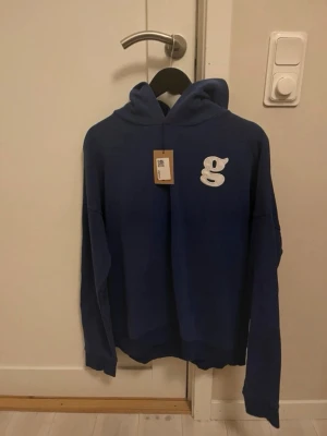 Blå hoodie med broderad g-logga - Fin blå hoodie med en broderad vit g-logga på bröstet. Tröjan har huva, långa ärmar och en relaxed passform. Perfekt för dig som gillar streetwear och vill ha något bekvämt och stilrent. Det är storlek large men skulle säga att den sitter mer som en medium. Ingen aning vilket märke det är!