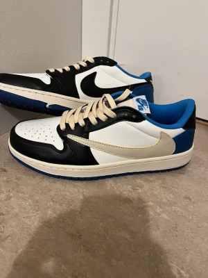 Nike Air Jordan 1 Low Travis Scott - Nike Air Jordan 1 Low Travis Scott i svart, vit och blå med beige snörning. Skorna har omvänd Swoosh, blå foder och detaljer som 'Cactus Jack' broderat på hälen. Tillverkade i skinn med perforerad tå och platt blå sula. Perfekt för dig som vill sticka ut med hypade sneakers.