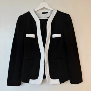 Svart och vit kavaj från XXFS - Stilren svart kavaj med kontrasterande vita detaljer från XXFS. Kavajen har vit kant längs öppningen och vita ficklock upptill, vilket ger en cool och modern look. Materialet är strukturerat och känns bekvämt. Perfekt för dig som gillar klassisk stil med en twist.