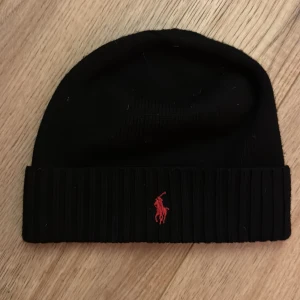 Svart mössa från Polo Ralph Lauren - Svart ribbstickad mössa från Polo Ralph Lauren med klassisk röd broderad logga framtill. Mössan har uppvikt kant och är tillverkad i mjukt stickat material som håller dig varm. Perfekt accessoar för kyliga dagar och snygg till streetwear. Knappt använd då ja inte passade i den. Lappen inuti är bortklippt då de kliade (se bild) 