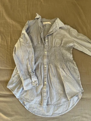 Blårandig skjorta med smock i midjan - Snygg blårandig skjorta från ZARA med smockdetalj i midjan som ger en cool siluett. Skjortan har långa ärmar, klassisk krage och knappar framtill. Tillverkad i mjuk bomull och passar perfekt för en avslappnad men trendig look.