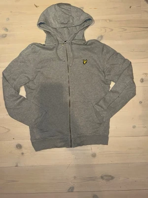 Grå zip hoodie från Lyle & Scott - Snygg grå hoodie från Lyle & Scott med dragkedja och huva. Klassisk gul logga på bröstet och justerbara snören i huvan. Tillverkad i mjukt bomullsmaterial som känns skönt mot huden. Perfekt för en avslappnad och stilren look. Men den har en lim fläck på ena armen men syns inte när man har den på sig