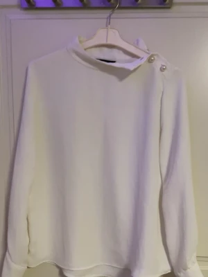 Vit blus med knappar vid kragen - Stilren vit blus med lång ärm och unik krage med två dekorativa knappar på axeln. Blusen har en rak passform och är tillverkad i ett mjukt, vävt material som ger ett elegant intryck. Perfekt för dig som gillar minimalistisk och modern stil.