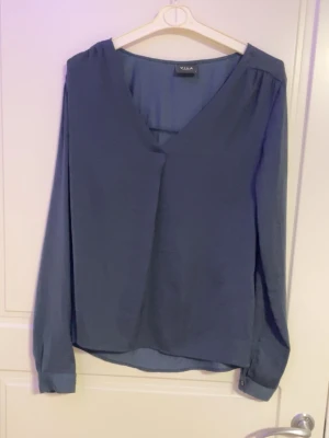 Blå långärmad blus från VILA - Stilren blå blus från VILA med v-ringning och långa ärmar. Tillverkad i ett mjukt, lätt tyg som ger ett snyggt fall. Perfekt till jeans eller kjol för en enkel men chic look. Klassisk design med knappdetaljer vid ärmsluten.