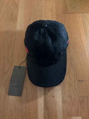 Svart keps från Gucci - Snygg svart keps från Gucci med diskret GG-monogram i tyget och klassiska gröna och röda ränder på sidan. Justerbar rem baktill och böjd skärm. Perfekt accessoar för att lyfta din outfit med lyxig känsla.