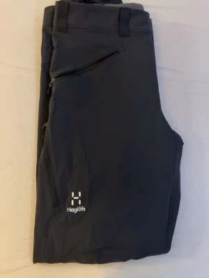 Haglöfs byxor svart - Säljer ett par svarta täckbyxor från Haglöfs med dragkedjor på fickorna och bälteshällor i midjan. Perfekta för vinterns alla äventyr. Snygg och funktionell design med diskret logga på benet.
