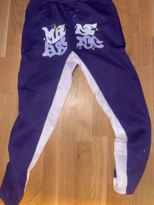 Lila mjukisbyxor med vit stripe - Säljer ett par lila mjukisbyxor med breda vita stripes längs benen och coolt tryck på baksidan. Byxorna har resår i midjan och är perfekta för chill dagar. Materialet känns mjukt och skönt, troligtvis bomull eller polyesterblandning.
