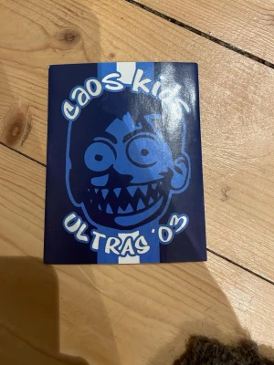 Caos Kids Ultras 03 klistermärke Dif  - Klistermärke med blå och vit bakgrund och en tecknad figur med spetsiga tänder. Texten 'Caos Kids Ultras 03' syns tydligt i vitt. Perfekt för att dekorera laptop, skateboard eller anteckningsbok.