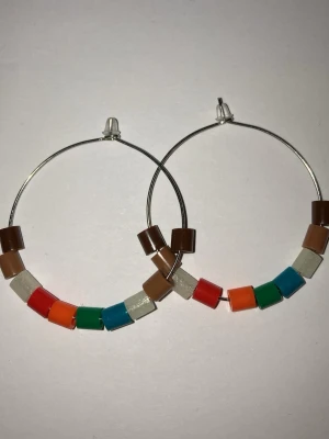 Färgglada hoops med pärlor - Stora hoops som är handgjorda av mig i silverfärgad metall med pärlor i olika färger som brunt, beige, rött, orange, grönt och blått. Och 4 av pärlorna lyser i mörkret. Pärlorna är kubformade. Perfekta för dig som gillar färg och unika accessoarer. Frakt 22-100kr beroende på vikten. Kontakta mig om du har frågor eller vill köpa.
