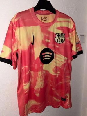 Barcelona x Nike matchtröja tie-dye - Unik Barcelona matchtröja från Nike med tie-dye mönster i rött och gult. Tröjan har klubbmärke, Nike-logga och Spotify som sponsor på bröstet. Tillverkad i lätt och ventilerande funktionsmaterial, perfekt för fotboll eller träning.