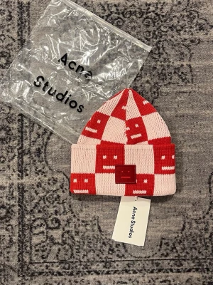 Röd och rosa mössa Acne Studios - Säljer en stickad mössa från Acne Studios i rött och ljusrosa rutmönster. Mössan har uppvikt kant och Acne Studios ikoniska ansiktsmärke framtill. Materialet är mjukt och stretchigt, perfekt för kalla dagar. Kommer med originalpåse och tagg.