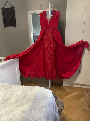 Röd långklänning med spetsdetaljer - Säljer en fantastisk röd långklänning med djup v-ringning och breda axelband. Klänningen har spetsdetaljer framtill, markerad midja och ett svepande, voluminöst kjolparti. Perfekt för bal eller festliga tillfällen. Materialet känns som polyester och spets.