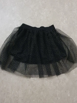 Kjol - Snygg svart kortkjol med glittriga guldprickar och flera lager tyll som ger extra volym. Resår i midjan för skön passform. Perfekt för dig som vill sticka ut lite extra med en trendig look.