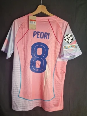 Barça matchtröja rosa Pedri Nike - Snygg rosa FC Barcelona matchtröja från Nike med Pedri och nummer 8 på ryggen. Tröjan har blå detaljer, Champions League-märke på ärmen och Spotify-logga på bröstet. Tillverkad i lätt och ventilerande material, perfekt för fotboll eller träning.