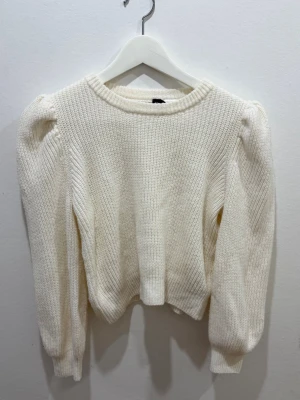 Vit stickad tröja  - Snygg vit stickad tröja  med puffärmar och rund halsringning. Tröjan har ribbade muddar vid ärmslut och nederkant, vilket ger en trendig och bekväm look. Perfekt att matcha med jeans eller kjol för en stilren outfit.