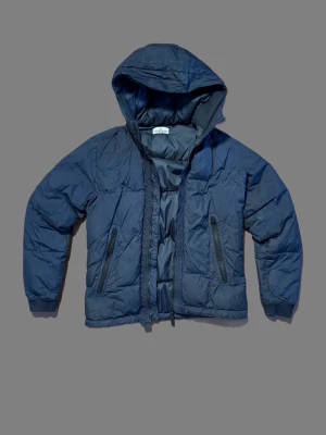Mörkblå Stone Island pufferjacka - Snygg mörkblå pufferjacka med huva och dragkedja framtill. Jackan har två praktiska fickor med dragkedja och en quiltad look. Perfekt för kyliga dagar och enkel att matcha med olika outfits.