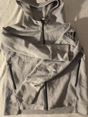 Adidas Hoodie - Ljusgrå hoodie från Adidas med dragkedja framtill och svarta detaljer. Stor Adidas-logga på ena ärmen och två fickor med dragkedja. Klassisk huva och mjukt material, perfekt för en avslappnad stil. Vid funderingar tveka inte på att höra av dig!