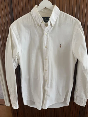 Vit skjorta från Ralph Lauren Slim Fit - Klassisk vit skjorta från Ralph Lauren i slim fit-modell. Skjortan har knappar framtill, button-down-krage och den ikoniska broderade loggan på bröstet. Tillverkad i mjuk bomull och har långa ärmar. Perfekt för en clean och stilren look.
