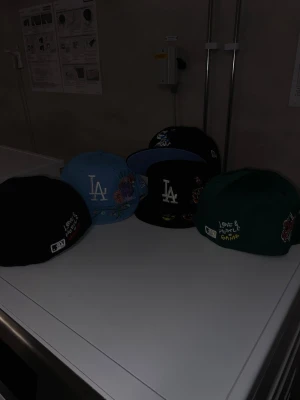 New Era fitted Cap Olika Design - Säljer flera LA Dodgers Fitted-kepsar från New Era i olika färger som svart, blå och grön. Kepsarna har broderade detaljer med blommor och texten 'Love & Hustle Game'. Klassisk platt skärm och justerbar passform. Perfekt för dig som gillar streetwear och snygga accessoarer.