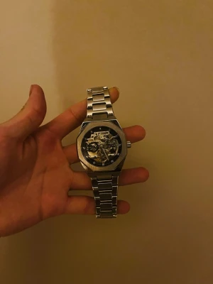 Silverfärgad skelettklocka i metall - Stilren klocka i silverfärgad metall med skeletturverk som visar de inre mekaniska delarna. Klockan har en robust länkarmband och en oktagonal boett. Perfekt för dig som gillar detaljer och industriell design.