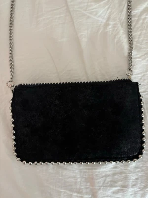 Svart axelväska med silverkedja - Snygg svart axelväska i mocka med silverfärgade kedjor och kuldetaljer runt kanterna. Väskan har dragkedja upptill och flera fack inuti för bra organisering. Perfekt storlek för det viktigaste och ger en cool vibe till din outfit.