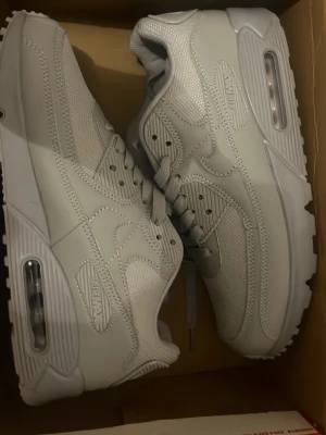 Nike Air Max 90 Surplus vita sneakers - Nike Air Max 90 Surplus sneakers i helvitt med klassisk chunky sula och synlig Air-enhet. Skorna har snörning, rund tå och är tillverkade i en mix av syntet och textil. Perfekta för dig som gillar stilrena och sportiga vibes. Helt nya