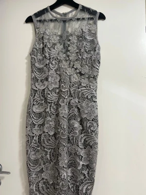 Grå spetsklänning med blommönster - Elegant grå klänning i spets med detaljerat blommönster och transparenta partier upptill. Ärmlös design och figurnära passform som ger en sofistikerad look. Perfekt för dig som vill sticka ut med en unik och stilren klänning.