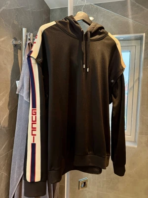 Svart hoodie från Gucci med ränder - Snygg svart hoodie från Gucci med huva och dragsko. Ärmarna har vita och blå ränder med röd GUCCI-text längs sidan, samt beige detaljer på axlarna. Tillverkad i mjukt material som känns lyxigt och bekvämt. Perfekt för dig som vill ha en exklusiv streetwear-look.