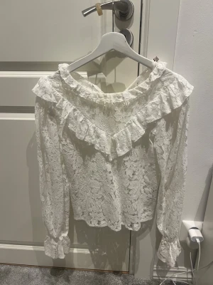 Vit spetsblus med volang - Romantisk vit blus i spets med långärmad design och bred volang runt axelpartiet. Blusen har en lös passform och fina detaljer vid ärmsluten. Perfekt för dig som gillar en feminin och trendig stil.