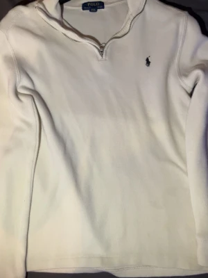 Vit halvzip från Polo Ralph Lauren - Klassisk vit tröja från Polo Ralph Lauren med halv dragkedja och broderad logga på bröstet. Tröjan har lång ärm och är tillverkad i ett mjukt, ribbat material som ger en clean och stilren look.