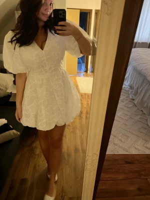Vit kort klänning med puffärmar - Superfin vit kort klänning med v-ringning, puffiga korta ärmar och markerad midja med knytband i ryggen. Klänningen har broderade blommönster och en lätt volangig kjol. Jag är 170cm och bär storlek L, klänningen går att justera i midjan efter ens kroppstyp pga knytbandet. Prislapp finns kvar, nypris: 899kr