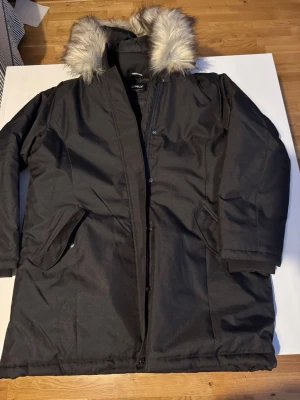 Svart parkas från ONLY med fuskpäls - Säljer en svart parkas från ONLY med stor huva och fluffig fuskpälsdetalj. Jackan har dragkedja och knappar framtill, två snedställda fickor och är lång i modellen. Perfekt för kalla dagar och riktigt snygg till vintern.