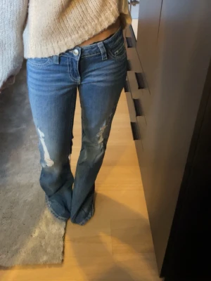 Blå bootcut jeans med slitningar - Säljer ett par blå lågmidjade bootcut jeans med snygga slitningar på benen. Jeansen är i fint skick med ytterst små slitningar längst ned.                                  Innerbenslängden är ca 76cm, midjemåttet är ca 36cm💓