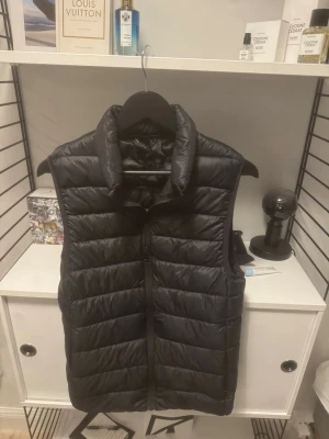 Svart dunväst med dragkedja - Säljer en svart dunväst med hög krage och horisontella quiltade sömmar. Västen har en stilren look och stängs med dragkedja framtill. Perfekt att slänga över en hoodie eller tröja för extra värme under kyliga dagar.