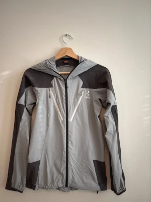 Grå vindjacka från Haglöfs - Snygg grå och svart vindjacka från Haglöfs med huva och hel dragkedja framtill. Jackan har två snedställda bröstfickor med vita dragkedjor och en sportig look. Perfekt för utomhusaktiviteter och äventyr.