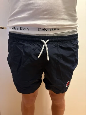 Champion Navy Blue Swim Shorts - Kids Size XXL (170/176) - Classic Champion swim shorts in a versatile navy blue. These feature white drawstrings and the iconic Champion logo on the left leg☺️  Size Note: These are Kids Size XXL (often corresponding to 170/176). As shown in the photos, they are too small for someone who is 180 cm tall. They would best fit a teenager or a shorter/slimmer adult looking for a very short fit☺️  The shorts are in excellent condition, made from high-quality quick-dry fabric, and have only been worn a few times☺️  