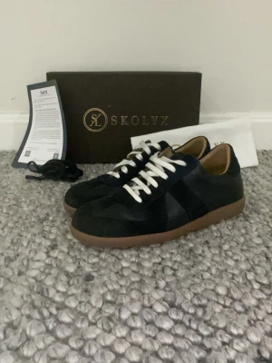 Blåa skolyx gast i mocka  - Snygga svarta sneakers från Skolyx gats i mocka med vita snören och bruna gummisulor. Klassisk låg modell med rund tå och stilren design som passar till det mesta. Levereras med extra skosnören och originalkartong. De till kommer även blåa snören. Sorry för dålig kavelitet. Skick nästa 10 av 10 använda ett fåtal gånger. Priset är inte hugget i sten och bara höra av sig vid funderingar😄