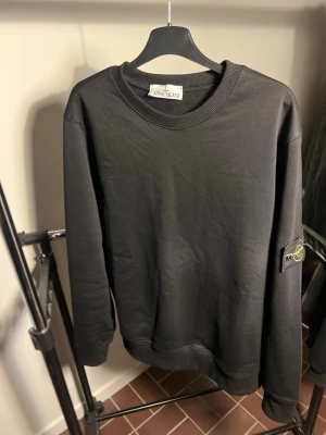 Stone Island - Svart sweatshirt från Stone Island med klassisk rund halsringning och ribbade muddar. Tröjan har den ikoniska Stone Island-loggan på vänster ärm och är tillverkad i mjukt bomullsmaterial. Perfekt för en avslappnad och stilren look.
