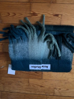 Grönmelerad halsduk Acne Studios - Säljer en fluffig halsduk från Acne Studios i grönmelerad ton med snygg färgskiftning från mörkgrönt till ljusare grönt. Halsduken har långa fransar i ändarna och en tydlig Acne Studios-logga. Perfekt accessoar för att lyfta din outfit.