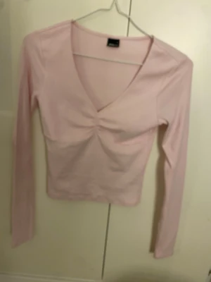 Ljusrosa långärmad topp med v-ringning - Söt ljusrosa topp med lång ärm och v-ringad hals. Toppen har en snygg rynkning framtill vid bysten och är figurnära i modellen. Perfekt att matcha med jeans eller kjol för en trendig look.