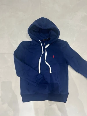 Blå hoodie från Polo Ralph Lauren - Mörkblå hoodie från Polo Ralph Lauren med vit dragsko i huvan och klassisk röd logga broderad på bröstet. Hoodien har känguruficka framtill och ribbade muddar vid ärmslut och nederkant. Perfekt för en avslappnad och stilren look.
