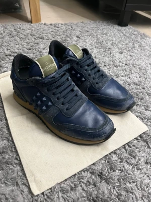 Valentino Rockrunner sneakers i marinblått skinn - Säljer ett par Valentino Rockrunner sneakers i marinblått skinn med svarta detaljer och ikoniska nitar på sidorna. Skorna har snörning, rund tå och platt sula. Kommer med original dustbag. Pris är inte hugget i sten och kom dm om du har några frågor 