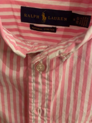 Rosa och vit randig skjorta Ralph lauren - Säljer en klassisk rosa och vit randig skjorta från Ralph Lauren. Skjortan har lång ärm, knappar framtill och en snygg krage. Tillverkad i bomull med natural stretch för extra komfort. Perfekt för en stilren och avslappnad look.