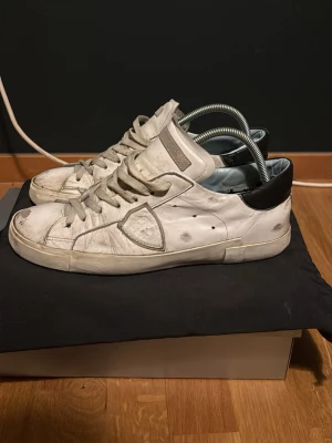 Vita sneakers från Philippe Model - Säljer ett par vita Philippe Model sneakers med svarta detaljer på hälen och grå snörning. Skorna har en klassisk låg siluett, rund tå och är tillverkade i skinn. Perfekta för dig som gillar stilrena och tidlösa sneakers. Rätt så slitna men har en lång användning kvar skriv för fler funderingar och priset kan diskuteras 
